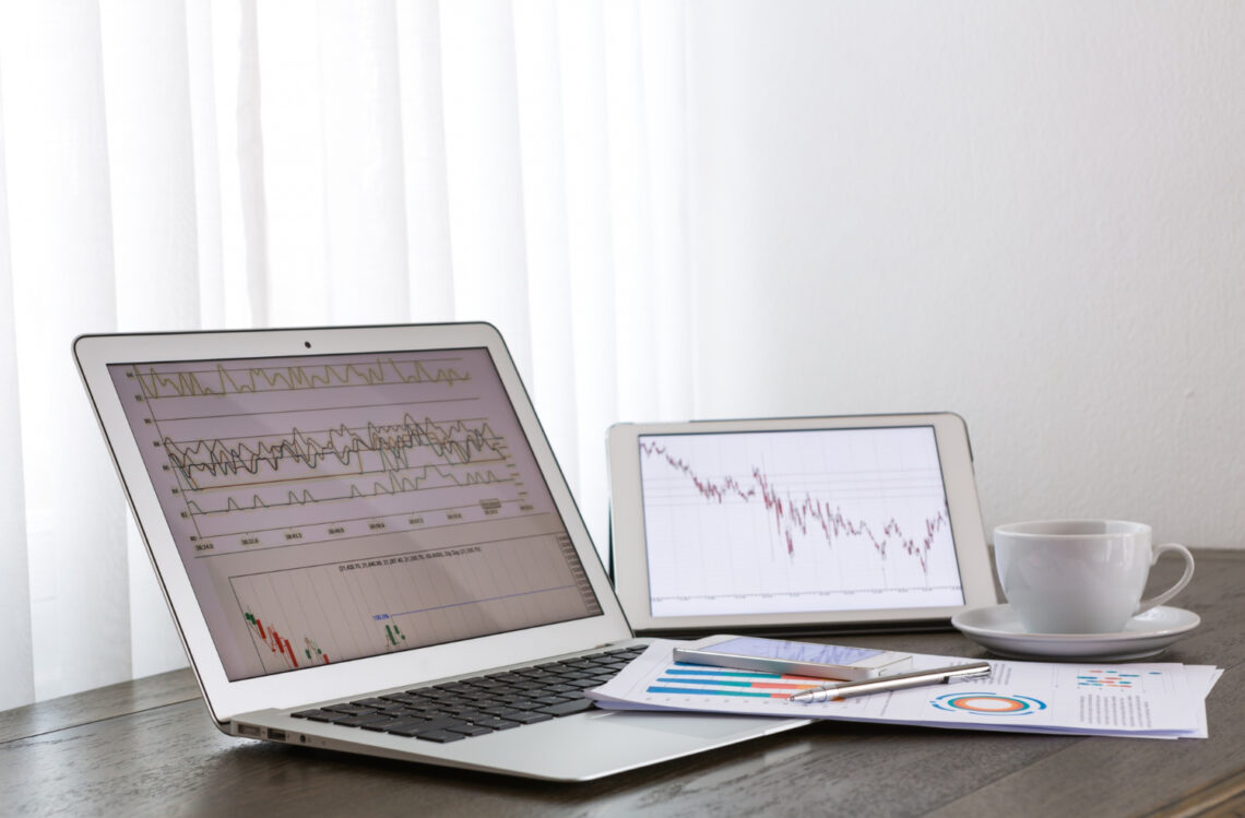 Open laptop and tablet displaying financial charts on a desk with documents, pen, and a cup of coffee beside them.