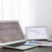 Open laptop and tablet displaying financial charts on a desk with documents, pen, and a cup of coffee beside them.